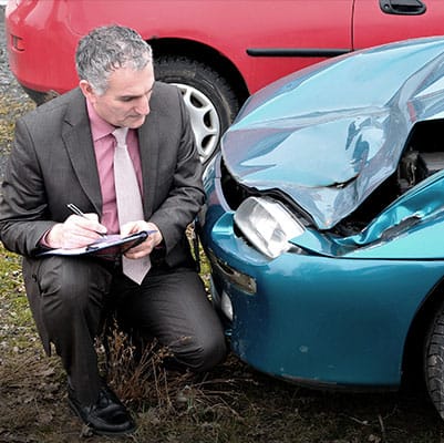 A man in a suit writes on a clipboard beside a damaged car, assessing the situation after an uninsured accident. - Brian Moore Injury Law